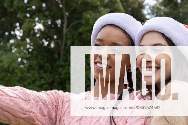 Diverse female friends posing for selfie in park, holding phone and wearing Santa hats, sweaters, model released, property released, 14.08.2025 06:00:00, 7811321