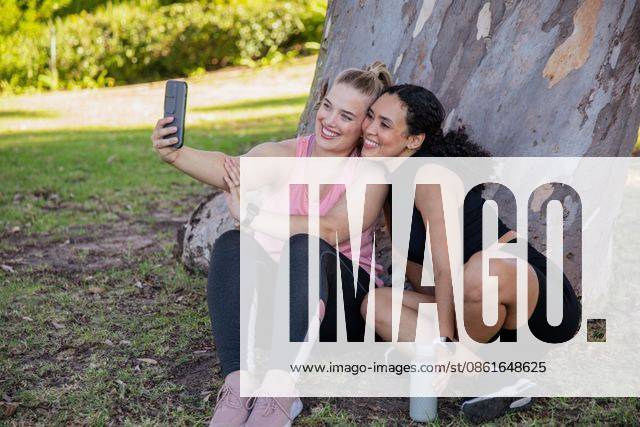 Diverse female friends posing for selfie while sitting on grass under tree in park using smartphone, model released, property released, 17.04.2025 06:00:00, 6702435