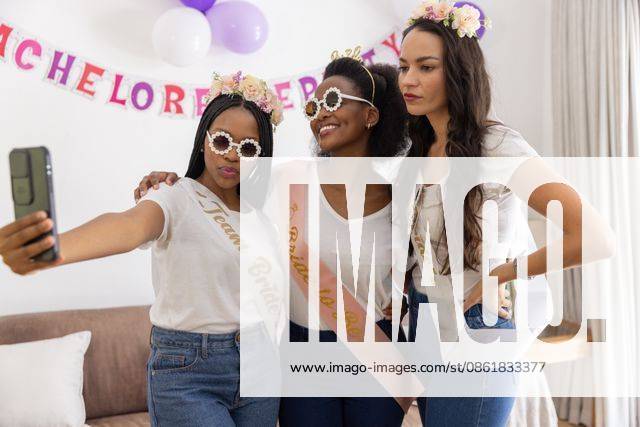 Diverse female friends posing for selfie at home with smartphone, banner, balloons, sashes, model released, property released, 29.11.2024 06:00:00, 6581250