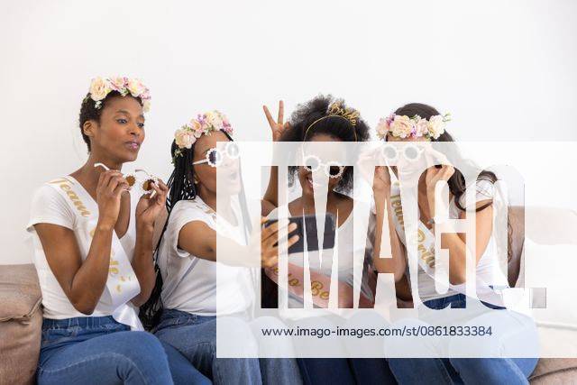 Diverse female friends posing for selfie on sofa wearing sashes sunglasses while holding smartphone, model released, property released, 29.11.2024 06:00:00, 6581265