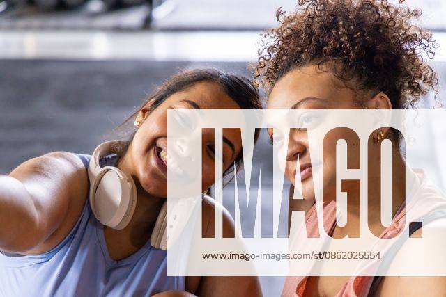 Diverse female friends posing for selfie in gym wearing athletic tops white headphones gold hoops, model released, property released, 19.08.2025 06:00:00, 7853528