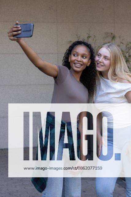 Diverse female friends posing for selfie, leaning toward smartphone by tiled wall with plaid shirt, model released, 30.10.2025 06:00:00, 9109305