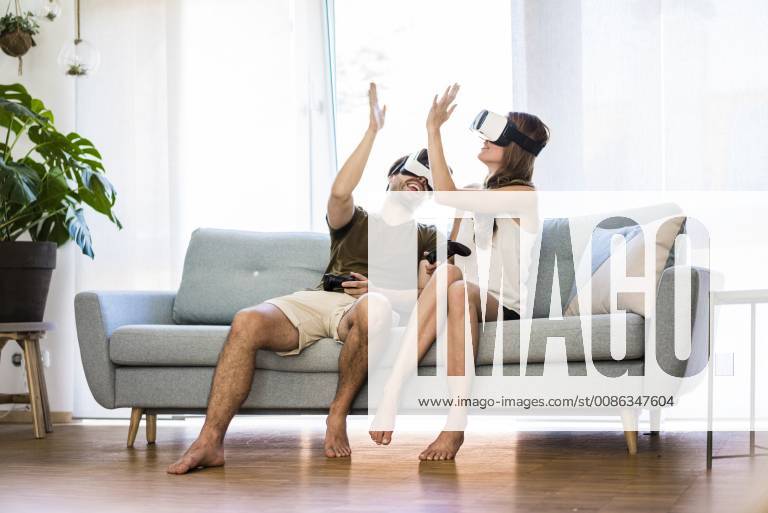 Happy couple sitting on couch at home wearing VR glasses high fiving model released Symbolfoto property released JOSF02566