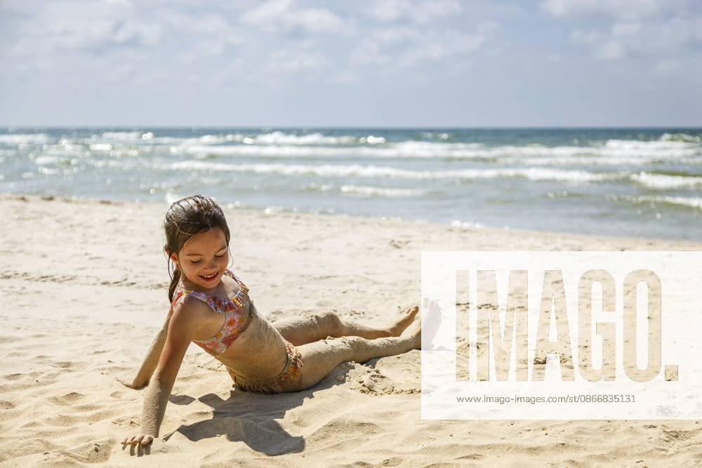 Ein süßes kleines Mädchen im Badeanzug sitzt am sandigen Strand, mit Sand bedeckt, während hinter ihr die Meereswellen brechen? ein Bild, das die Freude an den Sommerferien und unbeschwerte Kindheitserinnerungen verkörpert Ein süßes kleines Mädchen im Badeanzug sitzt am sandigen Strand, mit Sand bedeckt, während hinter ihr die Meereswellen brechen ? ein Bild, das die Freude an den Sommerferien und unbeschwerte Kindheitserinnerungen verkörpert