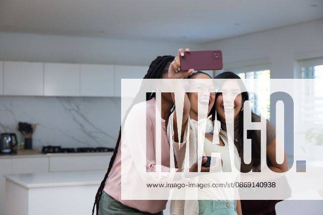 Diverse female friends posing for selfie in modern kitchen, holding red smartphone and headphones, model released, property released, 23.04.2026 06:00:00, 16412756