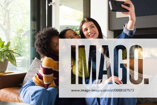 Diverse female friends posing for selfie on leather sofa in modern living room, holding smartphone, model released, property released, 21.05.2026 06:00:00, 16472232