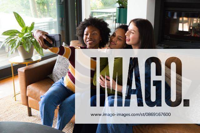 Diverse female friends posing for selfie on tan leather sofa in living room with smartphone, model released, property released, 21.05.2026 06:00:00, 16472399