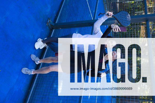 Diverse female friends posing for selfie on blue padel court holding black paddles, pink smartphone, model released, property released, 09.06.2026 06:00:00, 18050230
