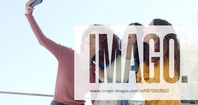 Diverse female friends posing for selfie on balcony holding phone by railing wearing denim jacket, model released, property released, Cape Town, South Africa, 02.09.2025 06:00:00, 10921366
