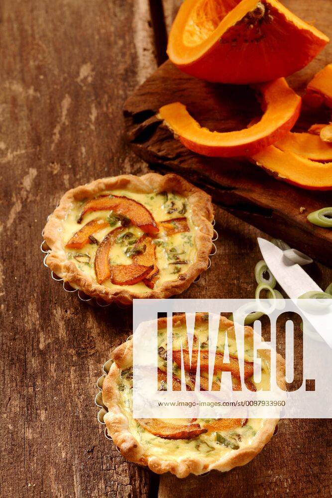 Appetizing small butternut, leek and custard quiches with a crispy pastry crust on a rustic wooden kitchen table with fresh ingredients, high angle view