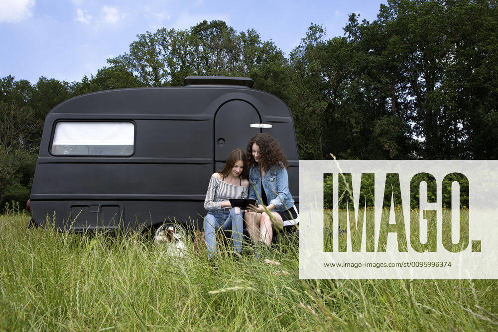 Two best friends sitting on a meadow in front of black caravan using digital tablet model released Symbolfoto property released PSTF00489
