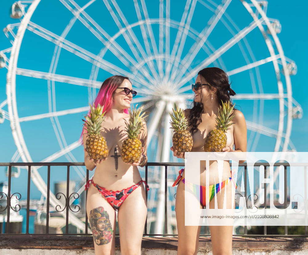 Young laughing women in bikini holding pineapples covering nude breast in  sunlight while laughing