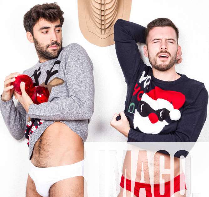 Couple of adult gay lovers in underwear and Xmas sweaters with decorative  red balls standing against white wall with artificial deer head stock photo