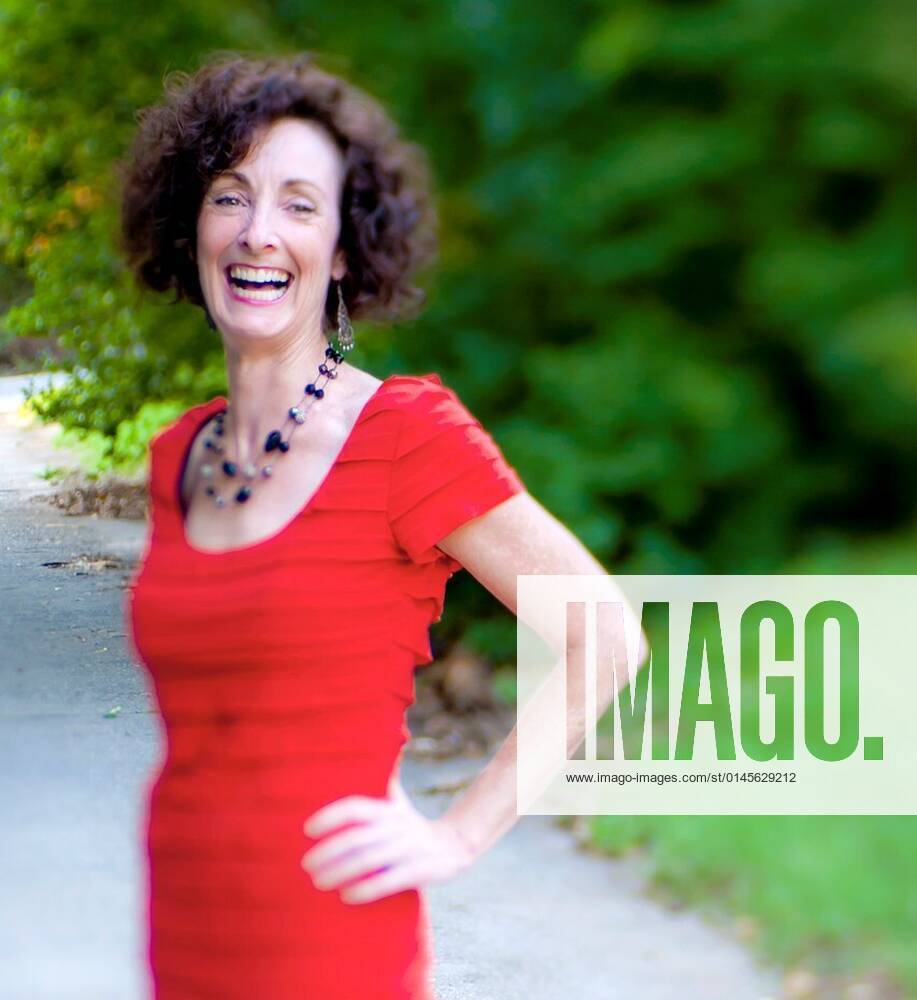 A 51 year old brunette woman wearing a red dress standing on a country  road. xagefotostockx