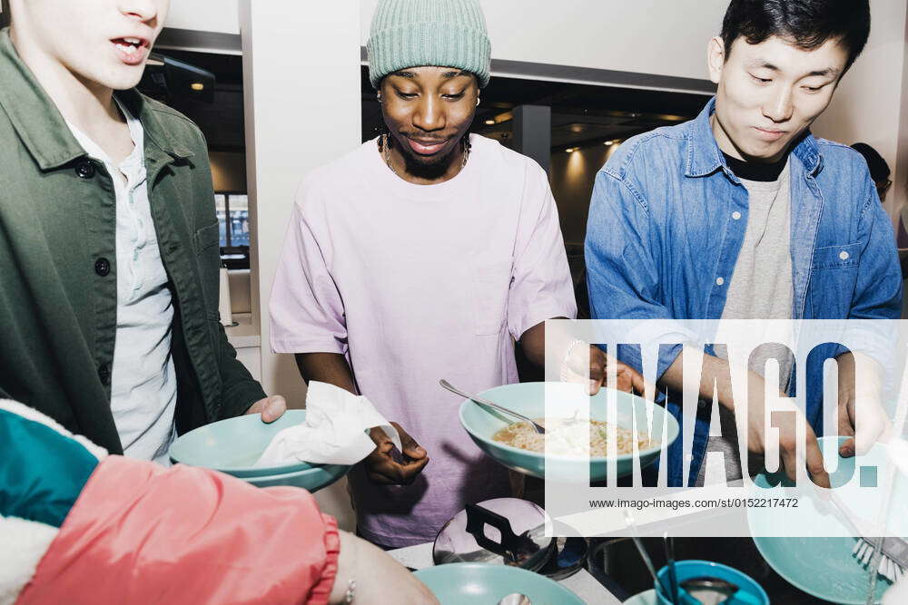 Young man with noodles while roommate cleaning dishes at sink in