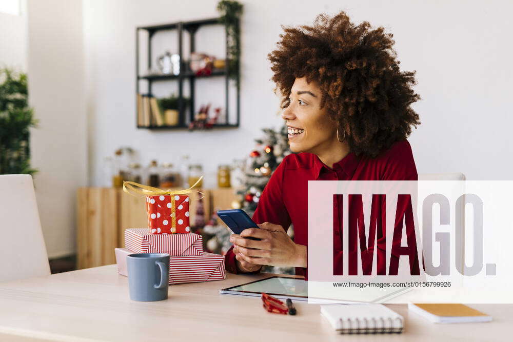 Happy Young Woman With Mobile Phone Sitting At Table In Living Room