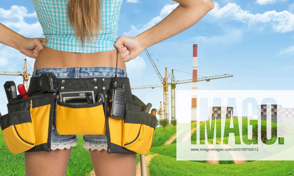 Woman in tool belt with different tools stands back. Hands on hip. Cropped image. Green hills and