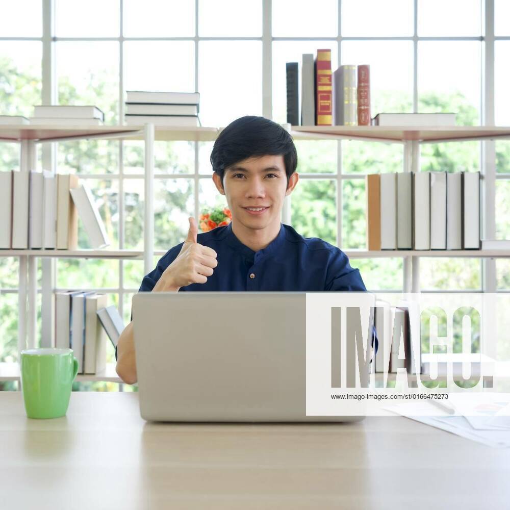 Young asian man lifts his thumb up with a smile behind laptop computer ...