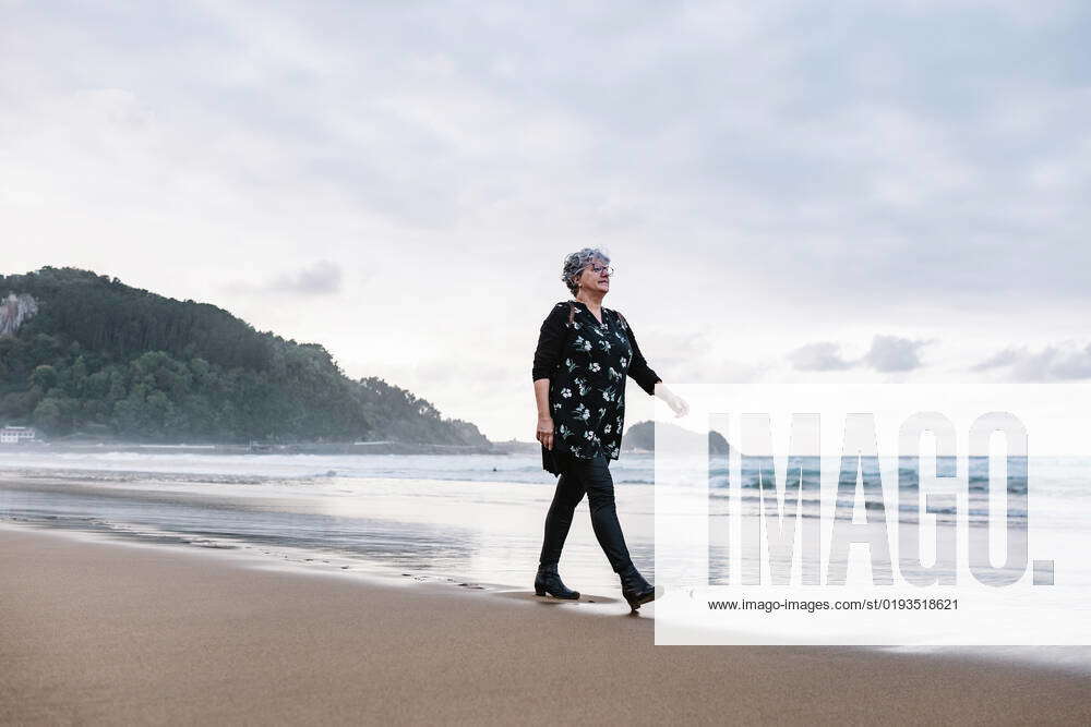 Full body elderly female traveler strolling on wet sand near waving sea ...