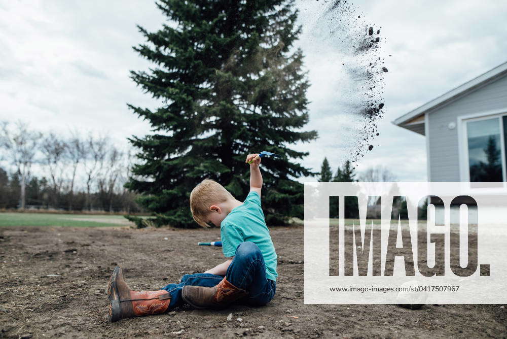 Side view of young boy sitting in yard while throwing handfuls of dirt ...