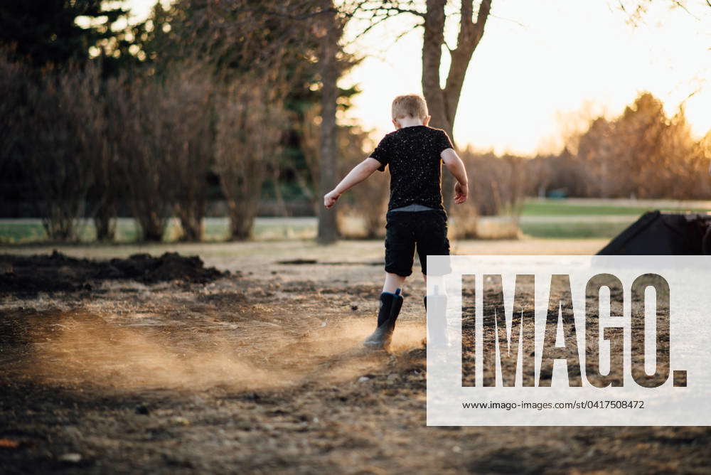 Wide read view of young boy kicking up dirt in yard on hazy summer eve ...