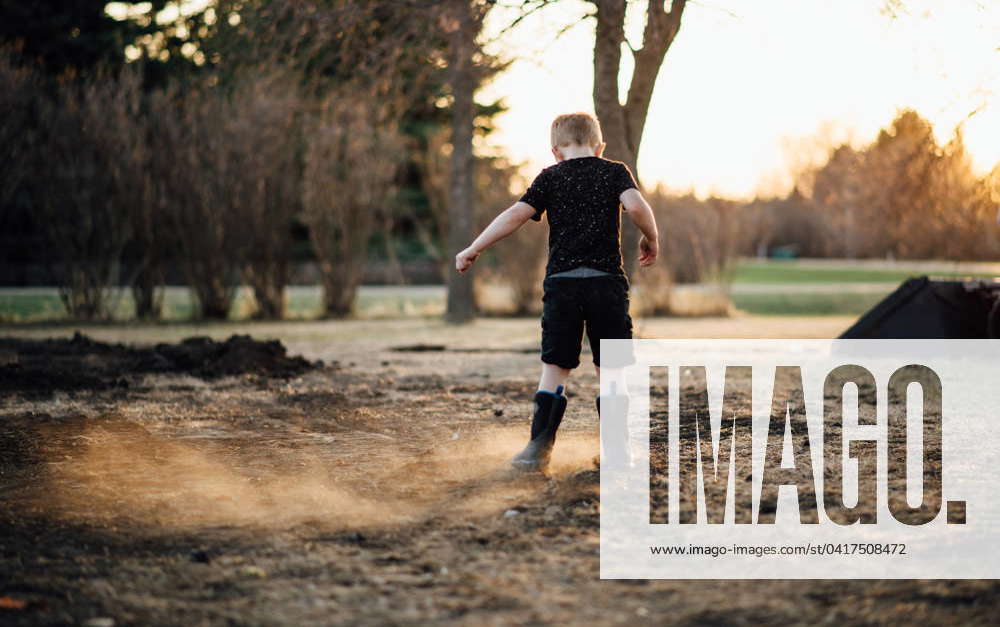 Wide read view of young boy kicking up dirt in yard on hazy summer eve ...